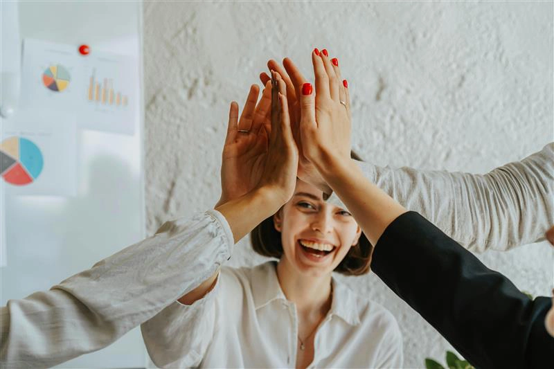 Mehrere Geschäftspersonen geben sich ein High-Five vor einem Whiteboard mit Diagrammen, symbolisiert Teamwork und erfolgreiche Zusammenarbeit im Büro.